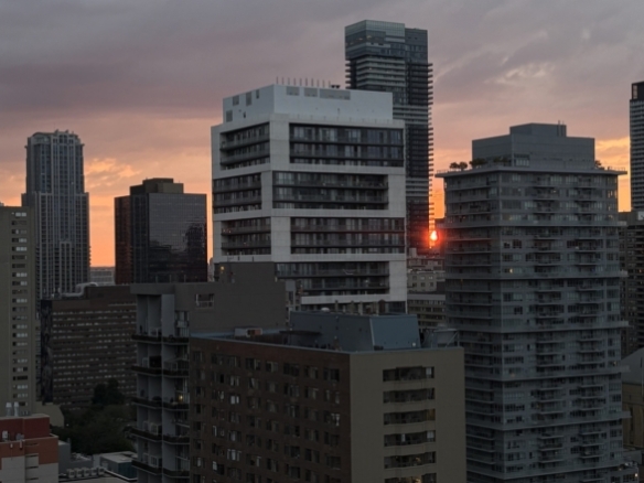 Image of a 7pm Toronto city skyline from the perspective of a 28th floor north-facing balcony window, during the summer. Shows a glowing pink sunset and the sun peeking through two condominium buildings.